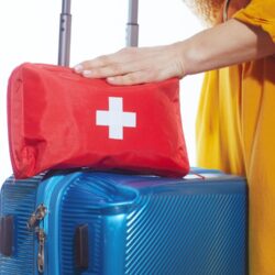 Červená lekárnička s bielym krížom leží na modrom kufri, vedľa ktorého stojí osoba v žltom oblečení pripravená na cestu.